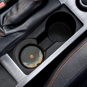 May include: A car cup holder with a round coaster featuring a celestial design. The coaster has a dark blue background with a yellow sun and white stars. The cup holder is black and silver, surrounded by black leather.
