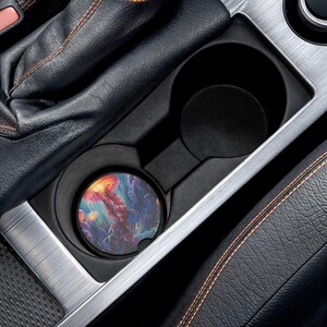 May include: A car cup holder with a colorful, round coaster featuring a vibrant jellyfish design. The coaster has shades of orange, red, purple, and blue. The cup holder is black and silver, set in a car's interior with black leather and orange stitching.