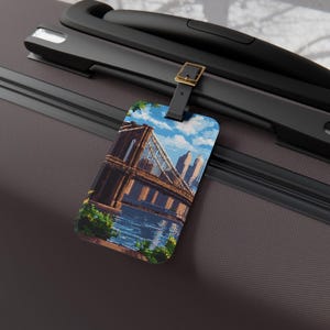 May include: A luggage tag featuring a pixelated image of the Brooklyn Bridge under a blue sky with clouds. The tag is attached to a black suitcase with a leather strap and a gold-colored buckle. The tag is rectangular with rounded corners.