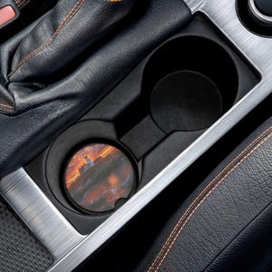May include: A round car coaster featuring a lighthouse scene at sunset. The coaster is placed in a car's cup holder, surrounded by black leather and silver trim. The artwork depicts a lighthouse on a cliff with a vibrant orange and red sky.