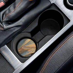 May include: A car cup holder with a coaster featuring a landscape design. The coaster has a circular shape and displays a scenic view with blue, brown, and yellow hues. The cup holder is black and sits within a silver trim.