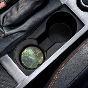 May include: A car cup holder with a forest-themed coaster. The coaster features a colorful landscape with trees and foliage. The cup holder is black and silver, and the car interior is black leather.