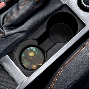 Könnte beinhalten: Ein runder Auto-Untersetzer mit einem Design des Big Ben-Uhrturms. Der Untersetzer befindet sich in einem Getränkehalter im Auto, umgeben von schwarzem Leder und silbernen Zierleisten. Das Zifferblatt der Uhr ist gelb und hat einen bunten Hintergrund.