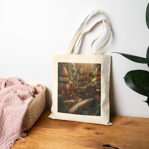 May include: A cream-colored tote bag displays a detailed illustration of a quaint village nestled in a valley. The artwork features charming houses, a stone bridge, and lush greenery, evoking a fairytale atmosphere. The bag is set against a neutral background.