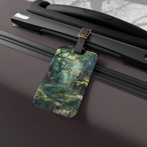 May include: A rectangular luggage tag with a detailed illustration of a fantasy forest scene. The tag has a black strap and a gold-coloured buckle. The artwork shows a path, trees, and small houses with a magical atmosphere.