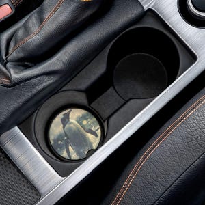 May include: A car cup holder coaster featuring a penguin illustration. The coaster is round with a black border and a snowy background. The coaster is placed inside a car's cup holder, surrounded by black leather and silver trim.