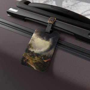 May include: Luggage tag featuring a landscape painting with a dramatic sky and a tornado. The tag is rectangular with a black strap and gold buckle, attached to a gray suitcase. The artwork depicts a rural scene with a storm.