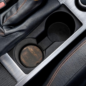 May include: A black car console cup holder with a wooden coaster. The coaster has a dark brown, weathered wood grain pattern. The console is surrounded by black leather and silver trim.
