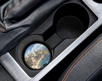 May include: A car cup holder with a coaster featuring a coastal town scene. The coaster has a detailed image of white buildings, blue water, and a bright sky. The cup holder is black and silver, set in a car's interior.