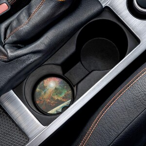 May include: A round car coaster featuring a colorful nebula design with shades of orange, green, and blue. The coaster is placed inside a car's cup holder, surrounded by black leather and silver trim.