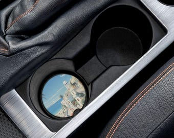 May include: A round car coaster featuring a coastal village scene with white buildings, blue skies, and ocean views. The coaster is placed in a black car cup holder, surrounded by black leather and silver trim.