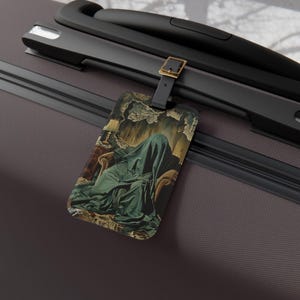 May include: Luggage tag with a surrealist painting of a figure covered in a green cloth, seated in a chair. The tag has a black strap with a gold-coloured buckle. The tag is on a dark grey suitcase.