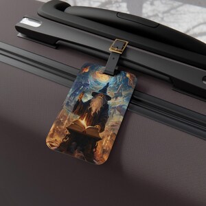 May include: A rectangular luggage tag featuring a wizard with a long white beard, wearing a blue hat and robe, reading from a glowing book. The background depicts a starry night sky with a full moon. The tag is attached to a black suitcase handle.