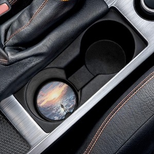 May include: A car cup holder with a round coaster featuring a person standing on a beach at sunset. The coaster has a colorful design with shades of orange, yellow, blue, and white. The cup holder is black and silver.
