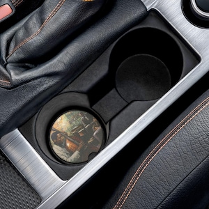 May include: A car cup holder with a round coaster featuring a painting of a jazz musician playing a cello. The coaster is in a black cup holder, surrounded by black leather and silver trim.