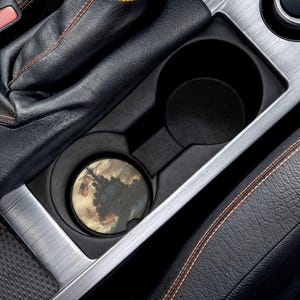 May include: A car cup holder with a coaster featuring a vintage illustration of a warship with an American flag. The coaster is round and fits snugly in the cup holder. The interior of the car is black with silver trim and orange stitching.