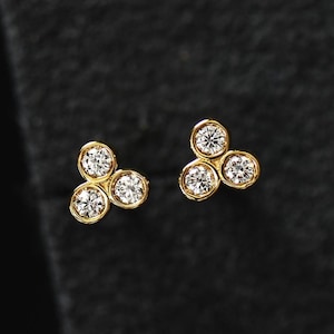 May include: A pair of gold stud earrings, each featuring three round, clear gemstones arranged in a cluster. The gemstones are set in a gold bezel setting, and the earrings are displayed against a dark background.