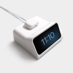 Peut inclure: Une station de charge blanche avec un chargeur sans fil blanc et une station d'accueil pour téléphone blanc. La station d'accueil pour téléphone a un écran noir affichant l'heure 11:10.
