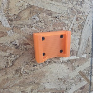 May include: An orange 3D-printed tool holder with four black screws. The holder is mounted on a wood-based panel. The holder has a rectangular shape with a curved bottom.