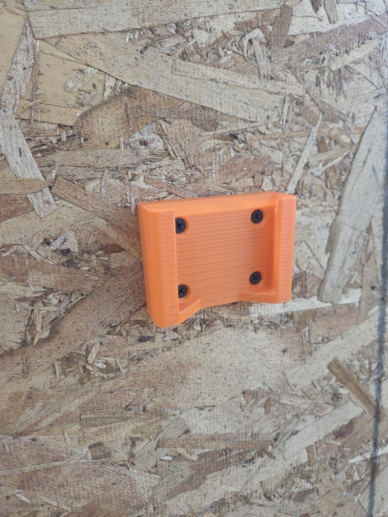 May include: An orange 3D-printed tool holder with four black screws. The holder is mounted on a wood-based panel. The holder has a rectangular shape with a curved bottom.
