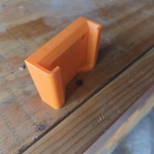 May include: An orange 3D-printed bracket with the text "II X II" on the top. The bracket is rectangular with a curved lip and is sitting on a wooden surface.