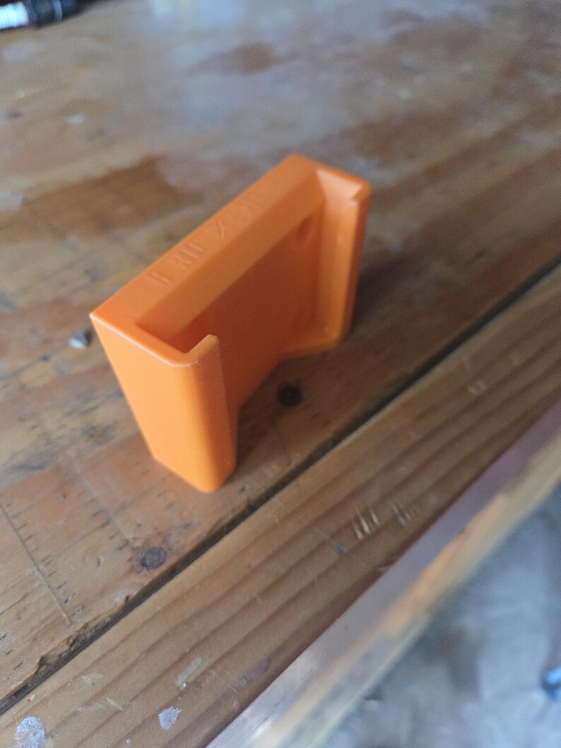 May include: An orange 3D-printed bracket with the text "II X II" on the top. The bracket is rectangular with a curved lip and is sitting on a wooden surface.