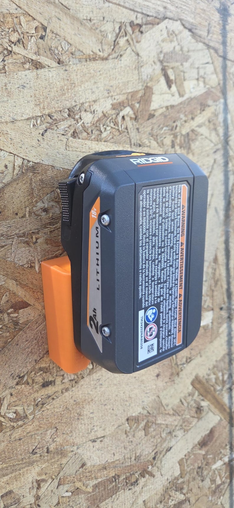 May include: A black and orange Ridgid 18V Lithium 2Ah battery pack for power tools. The battery is mounted on an orange holder. The image includes warning labels and the Ridgid logo.