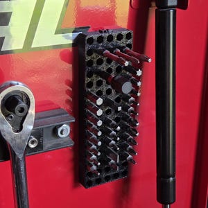 May include: A black, rectangular, 3D-printed tool organizer with a honeycomb pattern, mounted on a red metal surface. The organizer holds various metal screwdriver bits and hex keys in its hexagonal slots, arranged in rows.