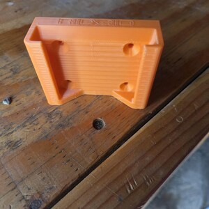 May include: An orange 3D-printed tool holder with the brand name "RIDGID" embossed on the top. The holder has a rectangular shape with angled sides and several circular holes for mounting. The item is sitting on a wooden surface.