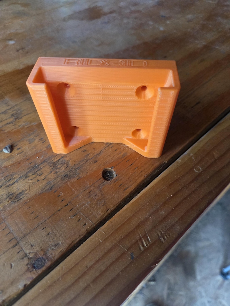 May include: An orange 3D-printed tool holder with the brand name "RIDGID" embossed on the top. The holder has a rectangular shape with angled sides and several circular holes for mounting. The item is sitting on a wooden surface.