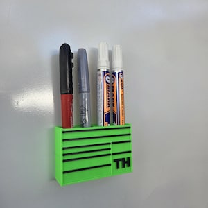 Könnte beinhalten: Ein limettengrüner Stifthalter in Form einer Werkzeugkiste, der vier Stifte hält. Die Stifte sind schwarz, silberfarben und zwei weiße Stifte mit blau-orangen Etiketten. Die Buchstaben "TH" sind auf der Vorderseite.