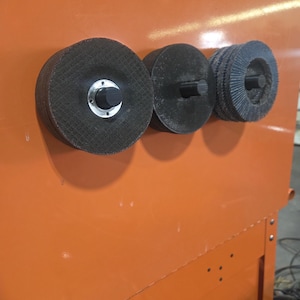 Puede incluir: Tres muelas abrasivas negras con orificios de montaje centrales están montadas sobre una superficie metálica naranja. Las muelas varían en diseño, una tiene un centro plateado y otra un borde estriado.