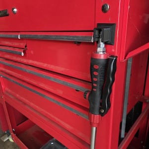 Puede incluir: Un cofre de herramientas de metal rojo con múltiples cajones y una pistola rociadora negra y roja adjunta. La pistola tiene un mango negro con detalles rojos y una boquilla plateada.