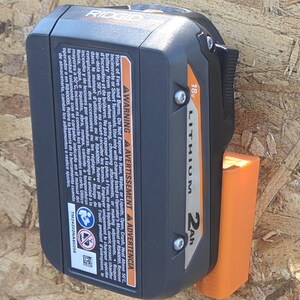 May include: Black and orange Ridgid 18V Lithium 2Ah battery. The battery is attached to an orange holder. The battery has a warning label and the text "LITHIUM 2Ah".