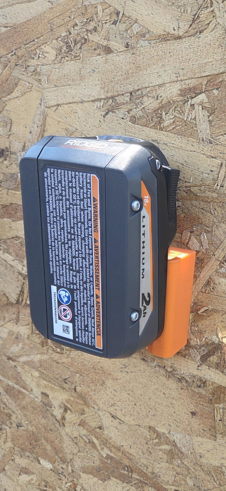 May include: Black and orange Ridgid 18V Lithium 2Ah battery. The battery is attached to an orange holder. The battery has a warning label and the text "LITHIUM 2Ah".