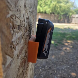 May include: An orange 3D-printed battery holder mounted on a wooden surface, holding a black and orange 18V Lithium 2Ah battery. The holder is designed to keep the battery secure and accessible. The background shows a green yard and blue sky.