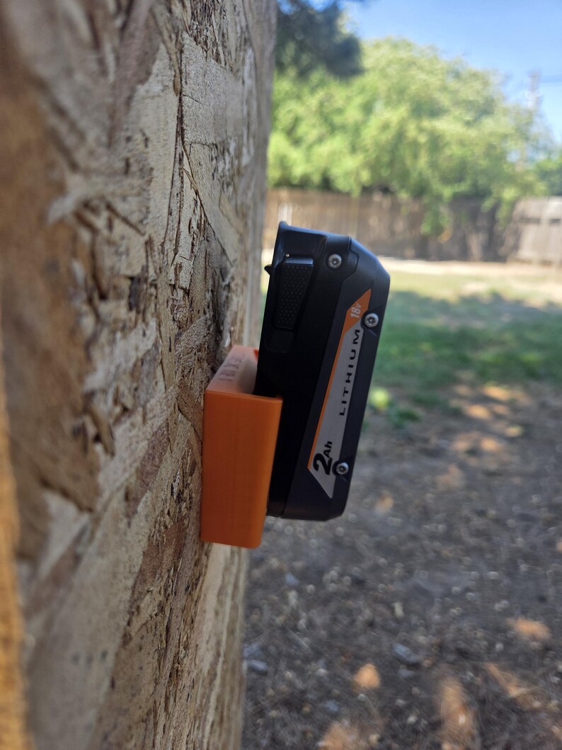 May include: An orange 3D-printed battery holder mounted on a wooden surface, holding a black and orange 18V Lithium 2Ah battery. The holder is designed to keep the battery secure and accessible. The background shows a green yard and blue sky.