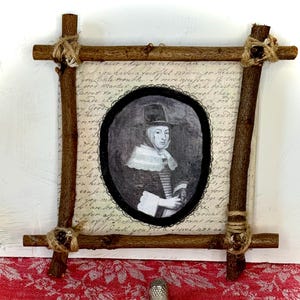 May include: A framed portrait of a person in a hat, set against a backdrop of handwritten script. The frame is crafted from rustic wooden branches, bound with twine. A silver thimble rests on a red and white patterned fabric.