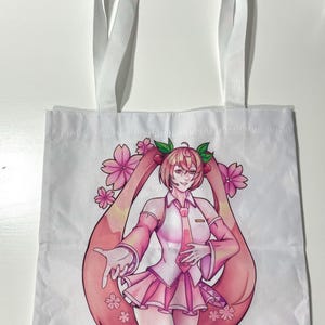 Puede incluir: Bolso tote blanco con un personaje rosa de estilo anime con pelo largo rosa y un atuendo rosa. El diseño incluye flores de cerezo rosas. El bolso tiene asas blancas.