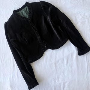May include: A black velvet bolero jacket featuring long sleeves with ruffled cuffs, a rounded front opening, and a ruffled trim along the neckline and front edges. The interior is lined with a solid olive green fabric, and the jacket has a cropped length.