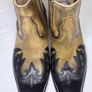 Könnte beinhalten: Ein Paar Stiefeletten im zweifarbigen Design mit einem Obermaterial aus metallisch goldfarbenem Leder in Used-Optik und einer glatten schwarzen Lederkappe. Die Stiefel haben dekorative schwarze Lederapplikationen mit Ziernähten und seitliche Einsätze.