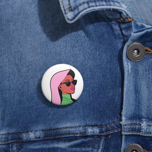 Op de afbeelding: Een witte knop met een roze en groene illustratie van een vrouw met een zonnebril. De vrouw heeft donker haar en draagt een roze hoofddoek en een groen shirt.