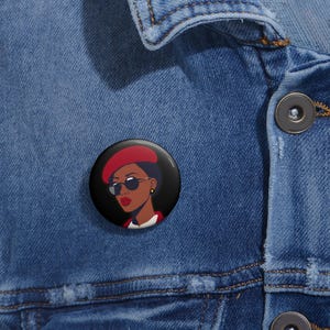 May include: A black and white button featuring a stylized illustration of a woman wearing a red beret and sunglasses.