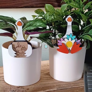 May include: Two white ceramic plant pots with decorative wooden goose stakes. One stake has a "smile" design, the other has colorful flowers. The stakes feature a white goose head with an orange beak and feet.