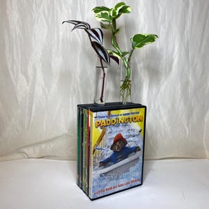 Puede incluir: Un soporte decorativo para plantas hecho de una pila de estuches de DVD, con la película "Paddington". Dos viales de vidrio contienen plantas verdes y moradas. El soporte está sobre una superficie blanca contra un fondo blanco.