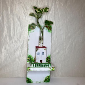 Könnte beinhalten: Ein handgefertigter Wandtopf aus Holz mit einem Miniaturhaus mit rotem Dach und weißen Wänden. Er enthält ein Glasfläschchen mit einer grün-weißen Pflanze. Das Design umfasst einen kleinen grünen Zaun und bemalte dekorative Akzente.