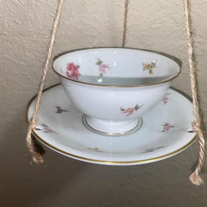 May include: A white teacup and saucer set with floral designs and gold trim, suspended by twine. The teacup sits atop the saucer, both adorned with pink, purple, and yellow flower patterns. The set is designed for decorative purposes.