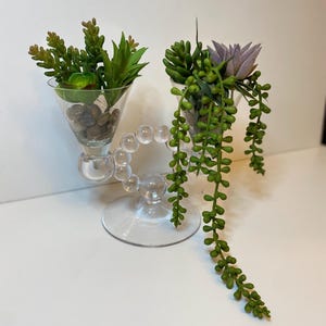 Może przedstawiać: Dwa przezroczyste szklane kieliszki do martini z dekoracyjnymi nóżkami, każdy wypełniony sztucznymi sukulentami i małymi kamieniami. Jeden kieliszek zawiera zielone i brązowe sukulenty, a drugi fioletowy kwiat i zwisające zielone rośliny.