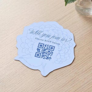 Może przedstawiać: Jasnoniebieska karta zaproszenia w kształcie kwiatu z tekstem "Will you join us? Please RSVP below" i kodem QR. Wazon z niebieskimi hortensjami stoi na jasnym drewnianym stole.