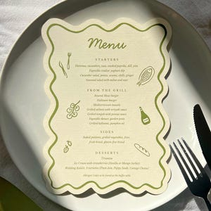May include: A menu card with a green wavy border sits on a white plate with a fork and knife. The menu lists starters, grilled items, sides, and desserts. The word "Menu" is at the top in a script font.
